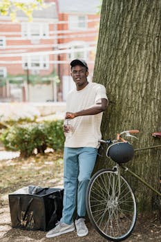 A young man stands with a bicycle and delivery bag, enjoying a break in the city park.