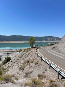 A scenic mountain road curves by a calm lake under a clear blue sky. Ideal for travel and nature themes.