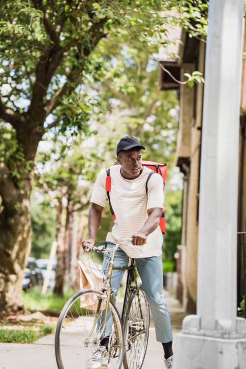 Free Afro-American courier riding a bike in a green urban setting delivering food on a sunny day. Stock Photo