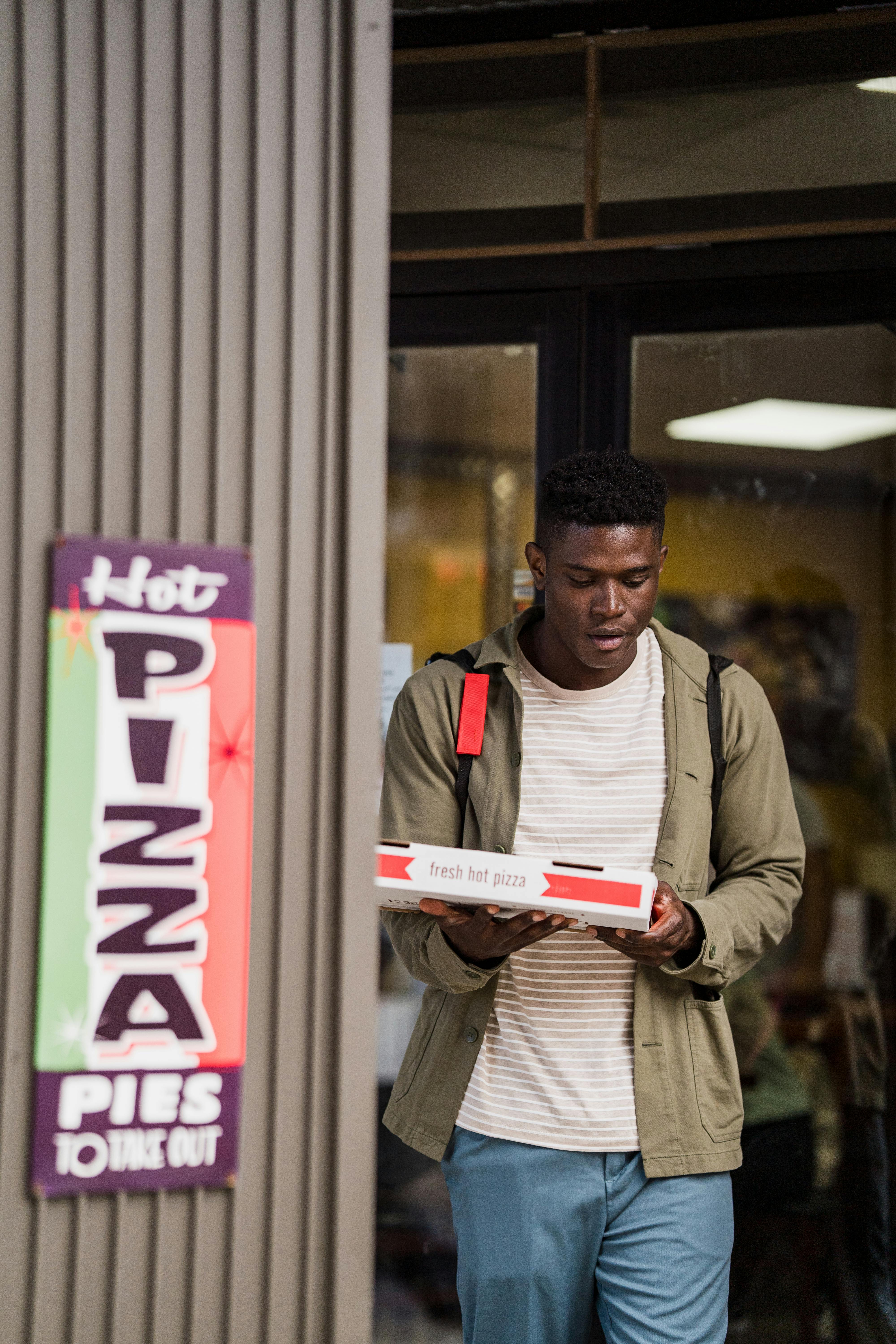 Man holding a pizza box outside a store, with signage indicating fresh hot pizza pies for takeout.