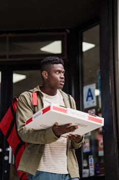 Young delivery person with a thermal bag carrying a pizza box outside a restaurant.