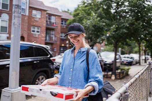 A cheerful woman in casual attire carries a pizza box in a city environment