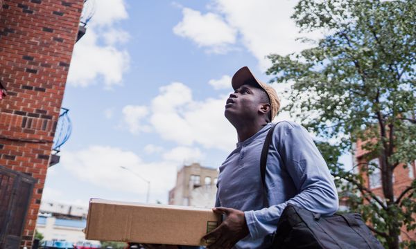 Last-Mile Delivery (Township) โ Man delivering a package in a city neighborhood, looking up at the building.