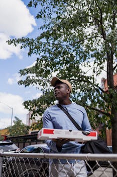 Young man in blue shirt carrying pizza box outdoors. Urban suburban backdrop with trees and buildings.