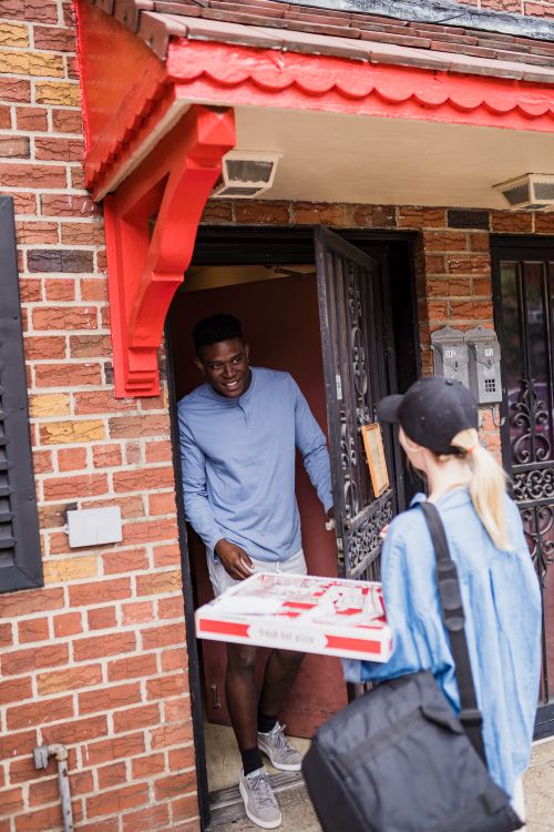 Free Smiling man receives pizza from delivery woman at brick house entrance. Stock Photo