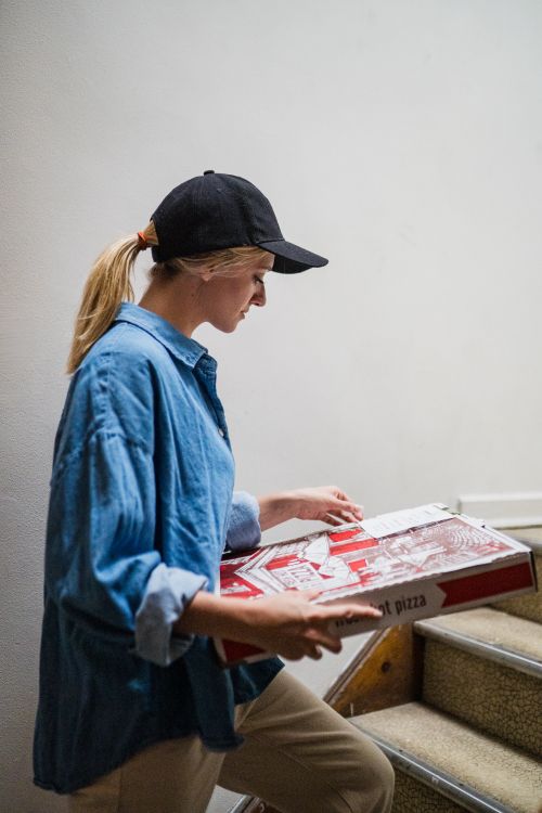 Free Young woman in casual attire carrying pizza boxes up a staircase indoors. Stock Photo