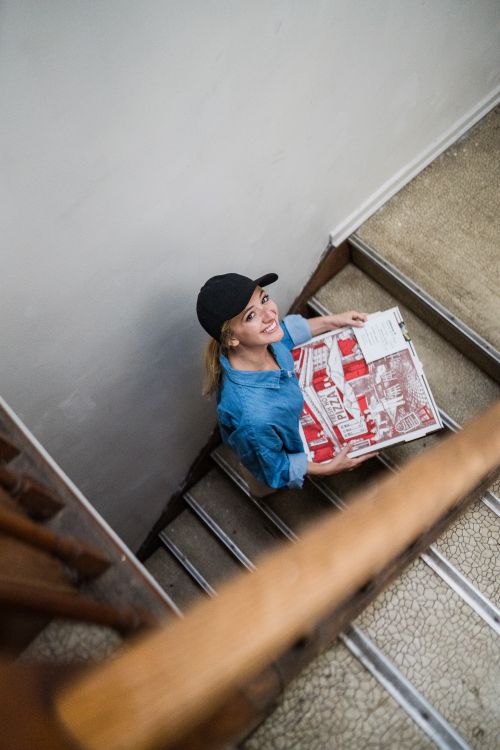 Free Smiling female courier carrying pizza boxes up a staircase indoors, viewed from above. Stock Photo