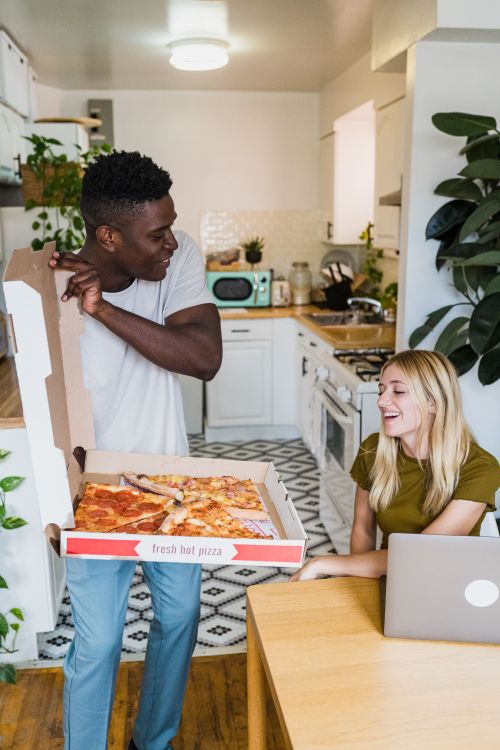 Free A cheerful couple sharing a pizza in a cozy kitchen setting with a laptop nearby. Stock Photo