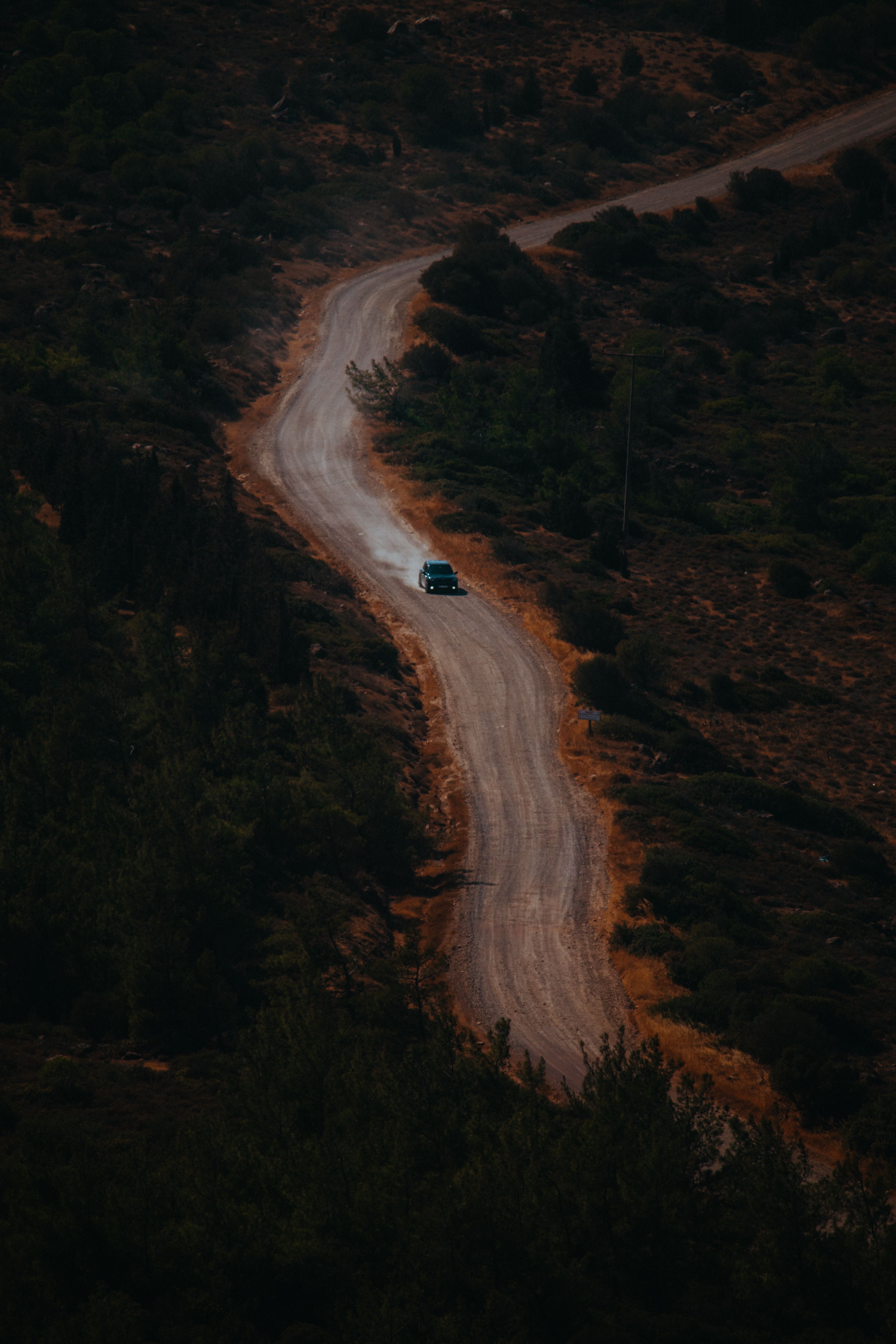 A car travels on a winding dirt road through a rugged forest area, captured from above.