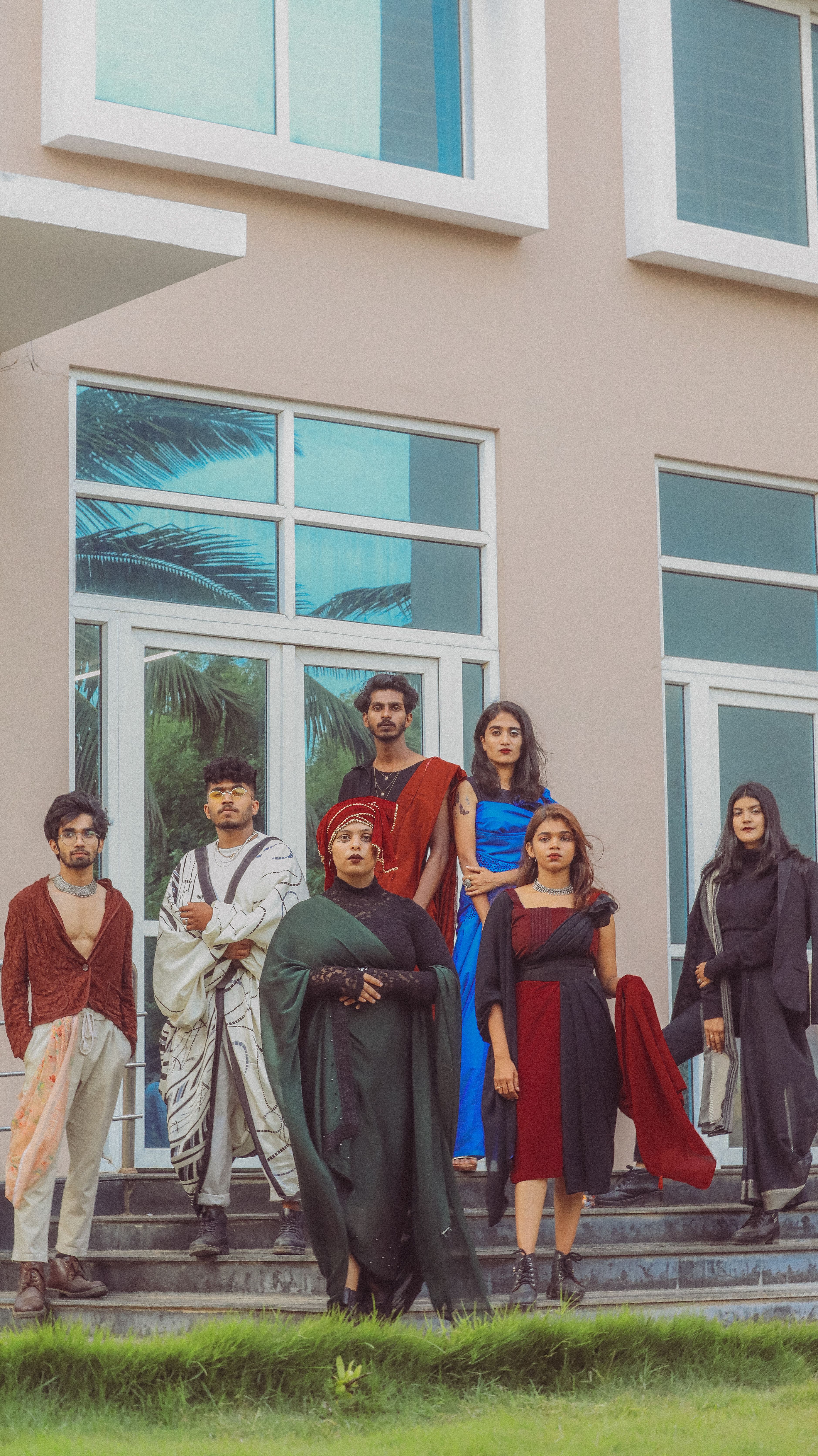 Seven people in traditional clothing posing by a modern building with glass windows.