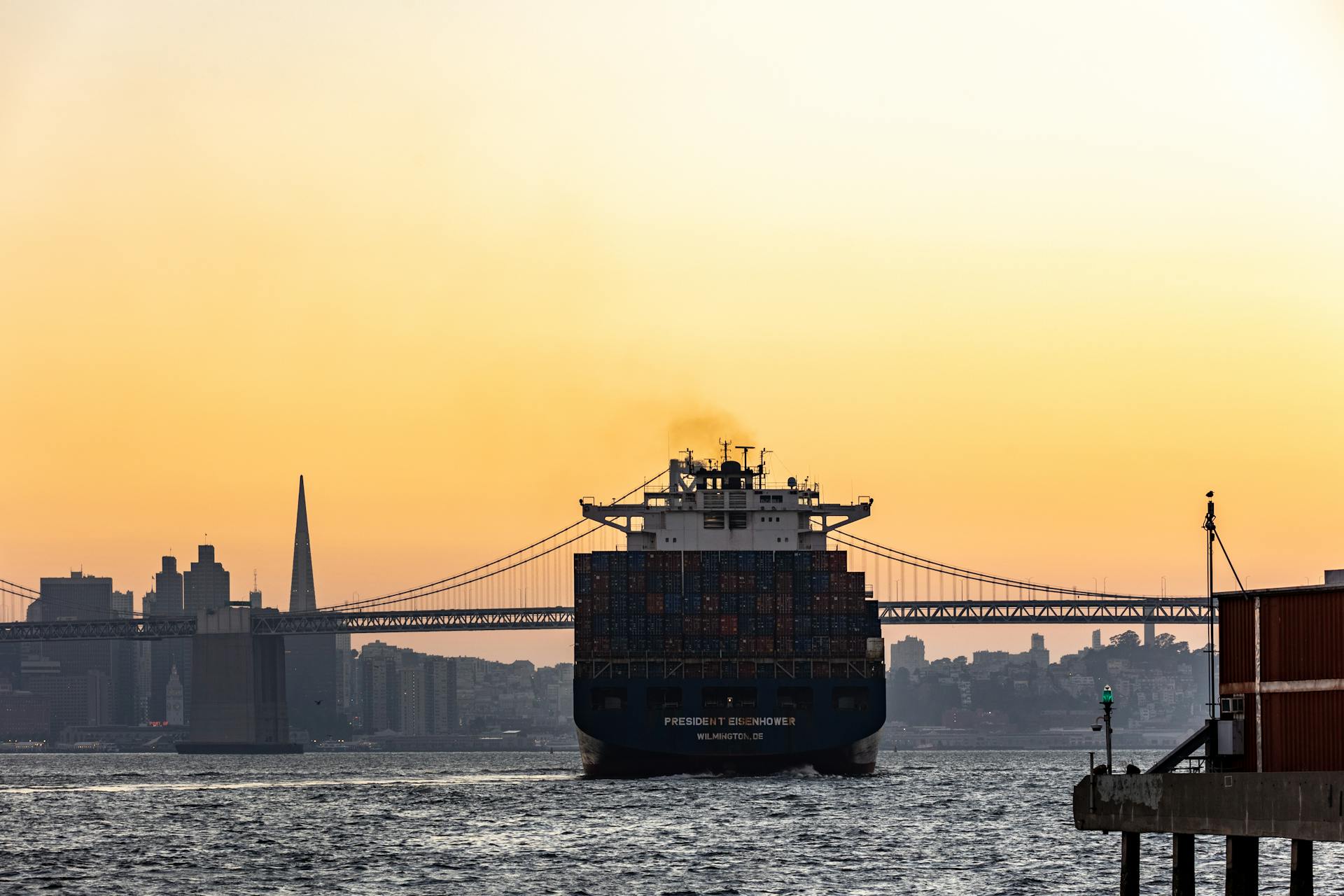 A container ship sails under a sunset sky near the San Francisco skyline and bridge, creating a stunning urban seascape.