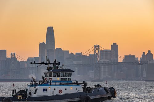 Free A tugboat navigates the waters with the San Francisco skyline and Bay Bridge at sunset in the background. Stock Photo