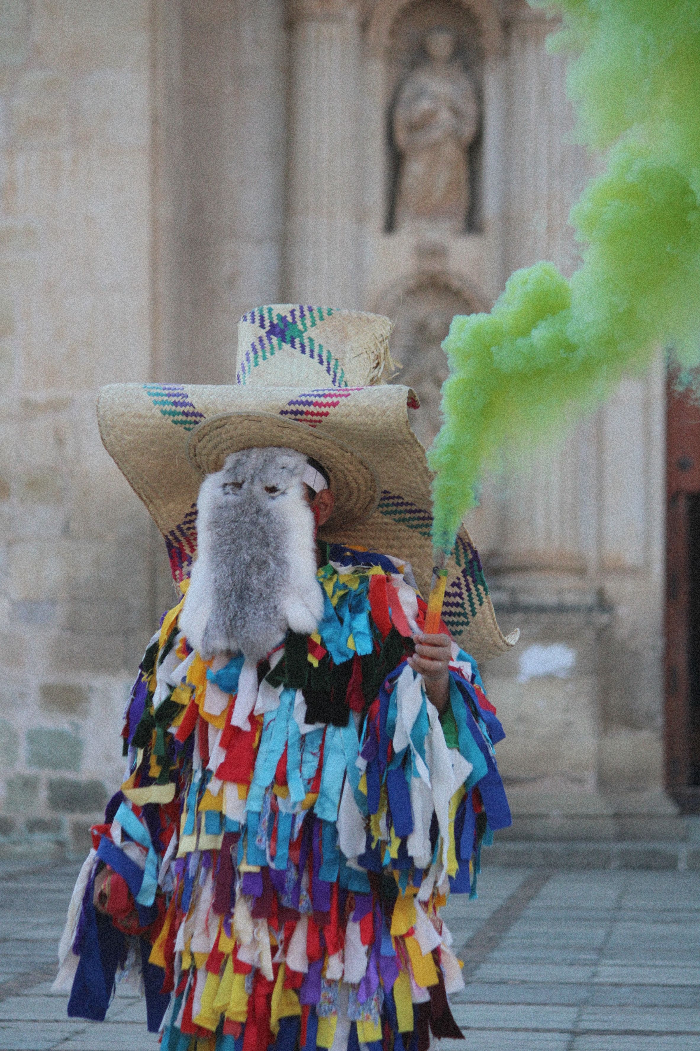 A person in a vibrant costume holding a green smoke flare in an urban setting.
