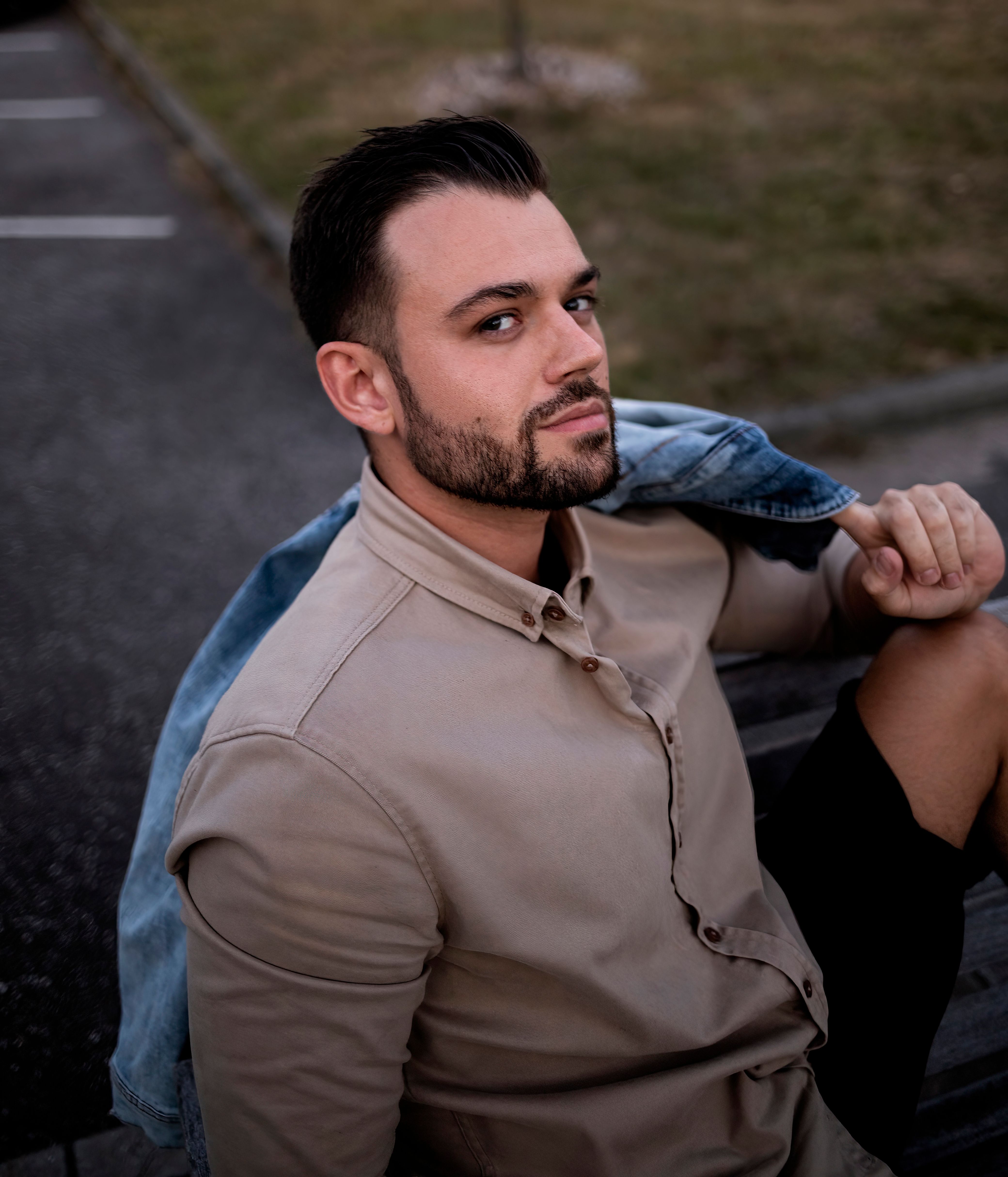 A bearded man outdoors, wearing a brown shirt and denim jacket, posing confidently.