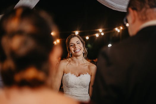 Smiling bride in elegant wedding dress outdoors at night with string lights.