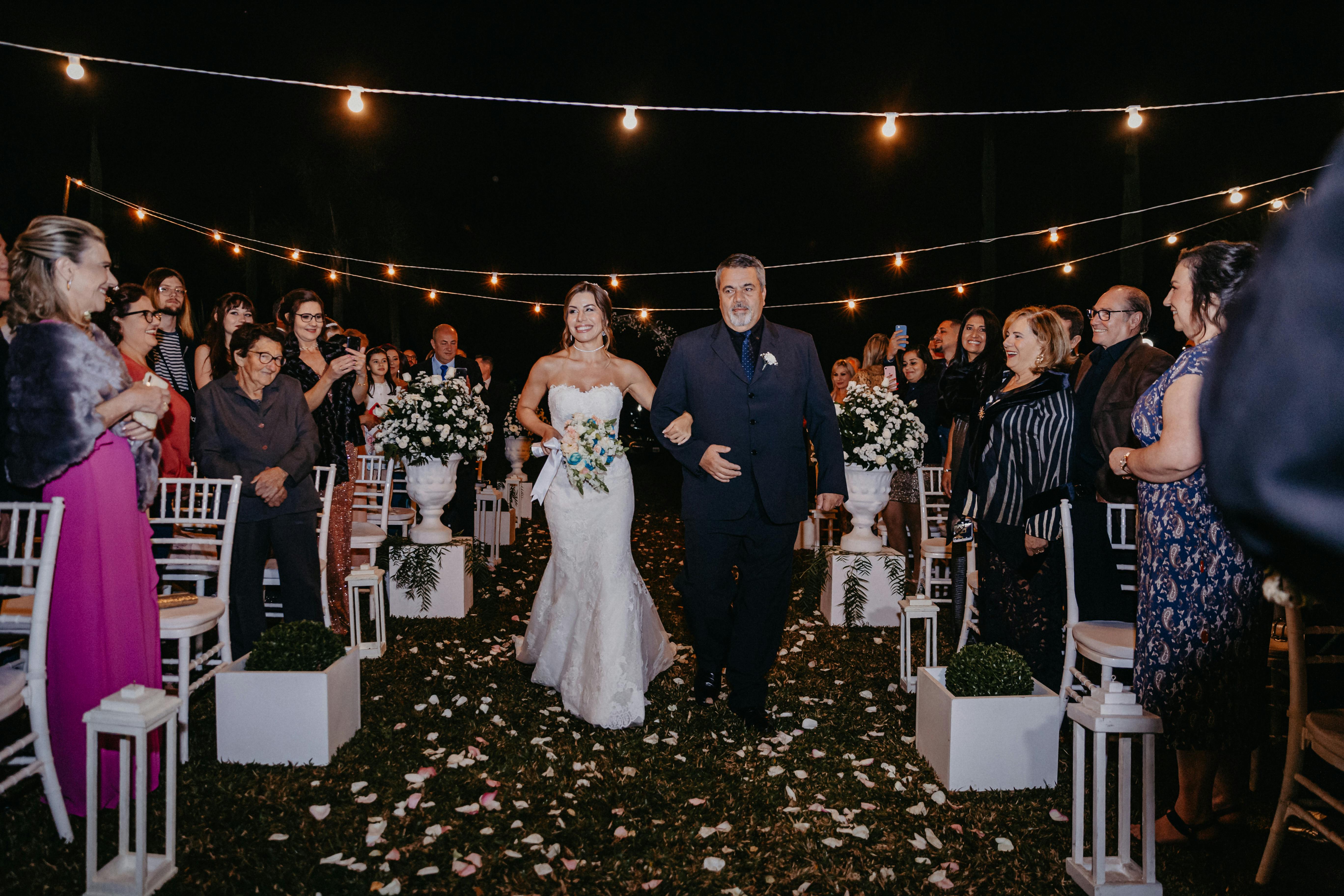 Bride walks down the aisle with her father at a beautiful outdoor night wedding, surrounded by guests.