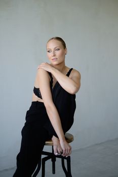A stylish woman in black attire posing gracefully on a stool against a neutral background.