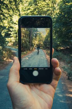A close-up of a smartphone capturing a scenic forest road with a cyclist.