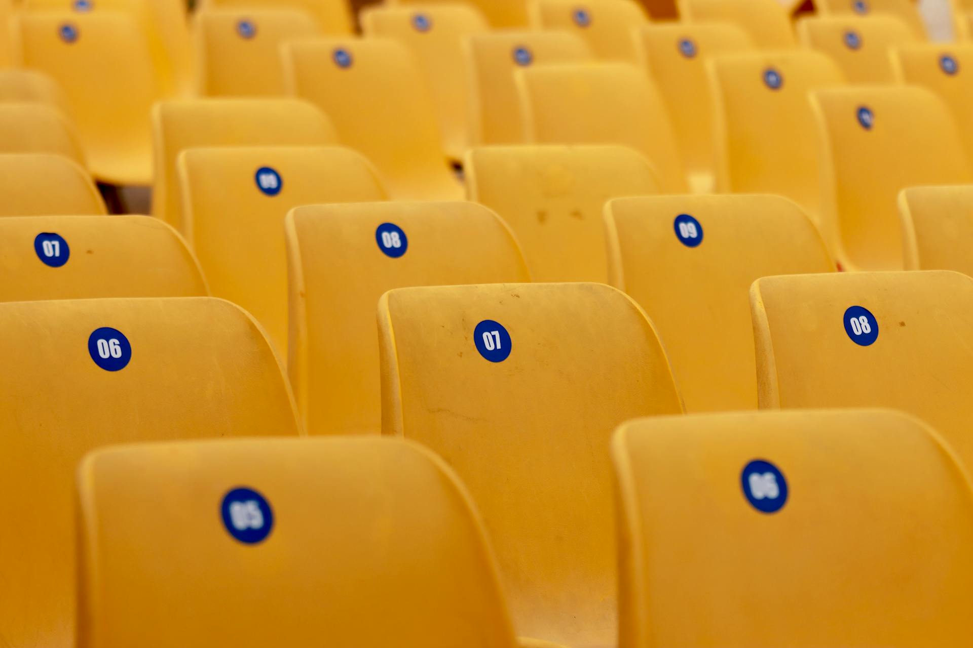 Rows of empty yellow plastic seats with numbered stickers in a stadium setting, offering a pattern and bright color.