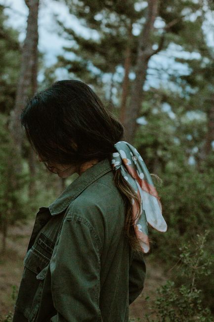 Young woman with scarf in hair stands amidst forest greenery in a serene setting.