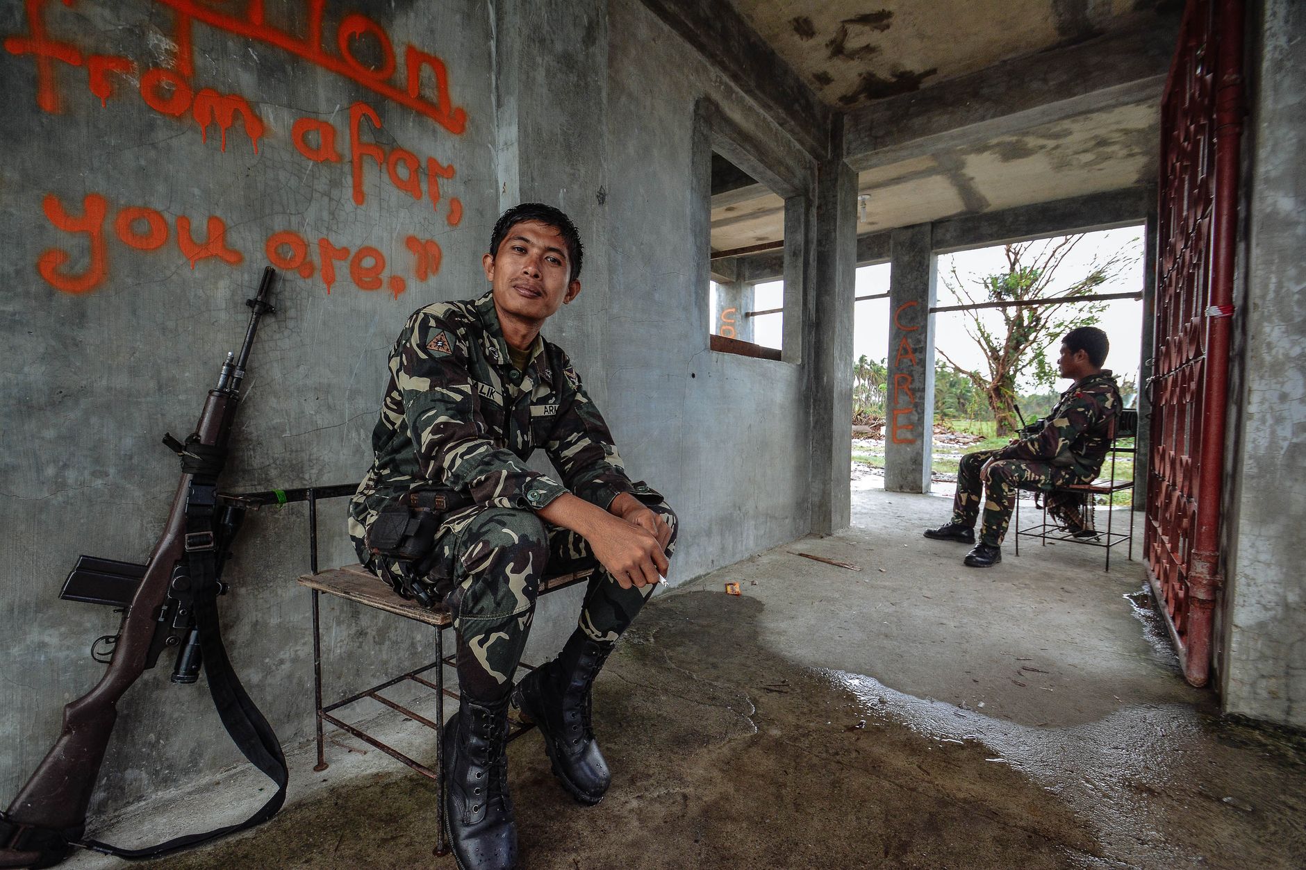 Two soldiers in camouflage uniforms sit inside an abandoned building with rifles nearby.