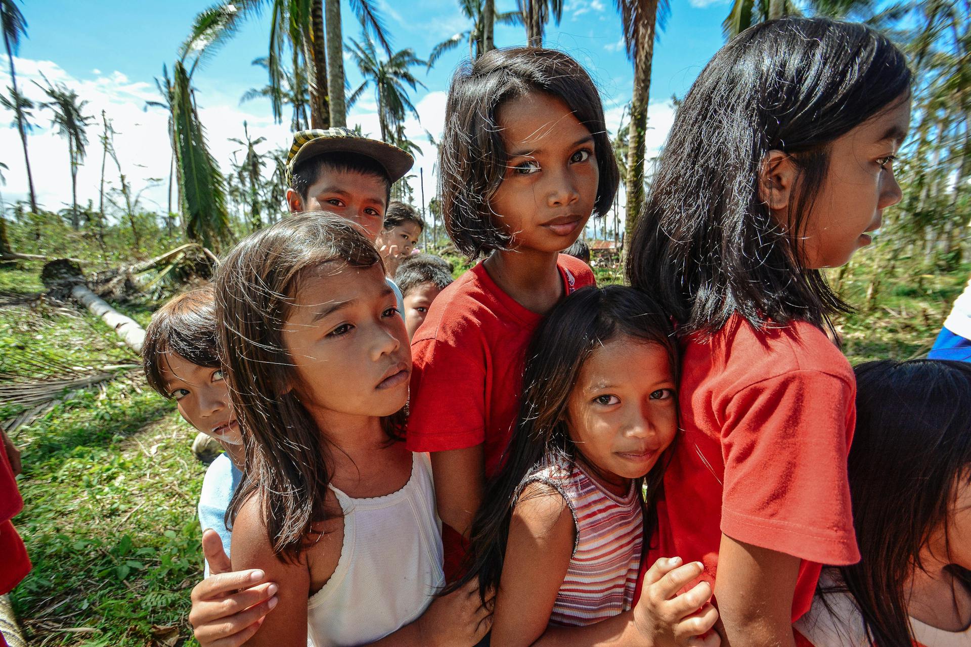 A diverse group of children stands together in a palm tree-lined area on a sunny day.