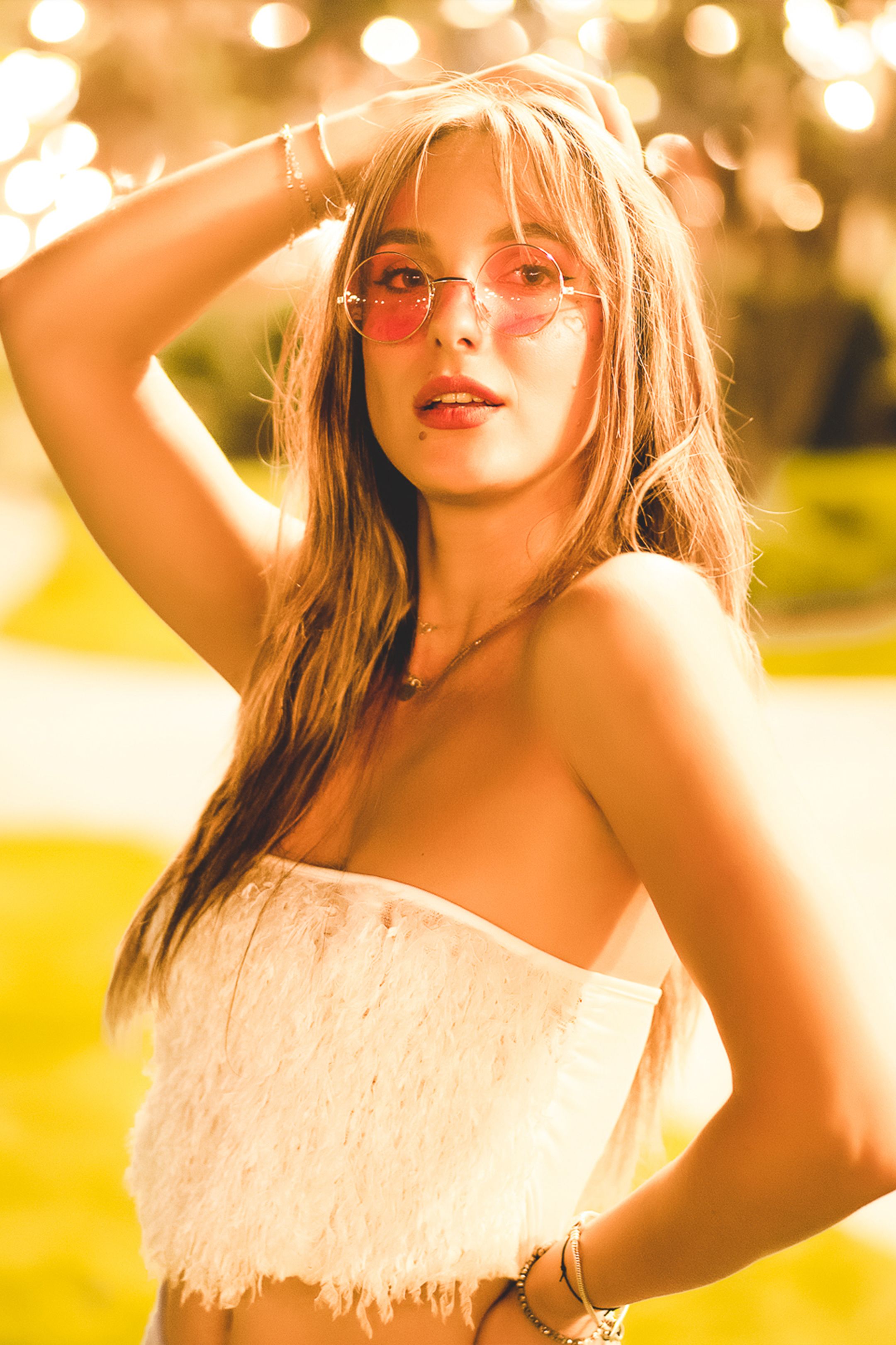 A stylish woman with long hair poses outdoors, wearing trendy sunglasses and a sleeveless top.