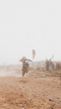 A motocross rider performing a jump on a dirt track amidst a dusty environment with spectators.