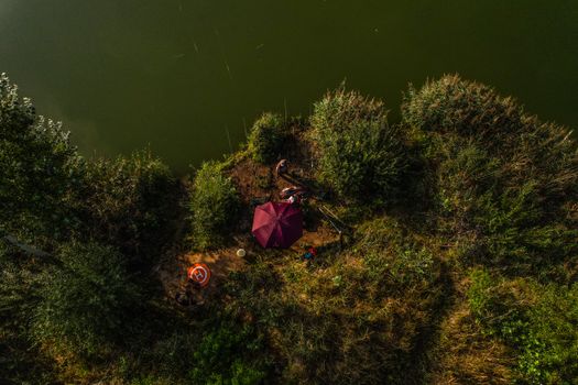 Drone shot of a lakeside campsite featuring fishing gear and a purple umbrella surrounded by lush greenery.
