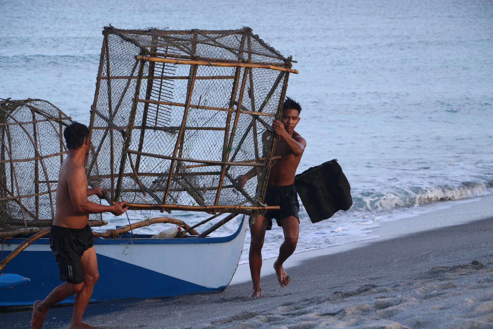 Two fishermen carrying a large fish trap on a beach, working early at dawn.