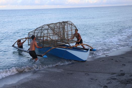 Three fishermen maneuver a traditional boat with fish trap on a sandy beach at sunrise. - Fotografer Sidoarjo