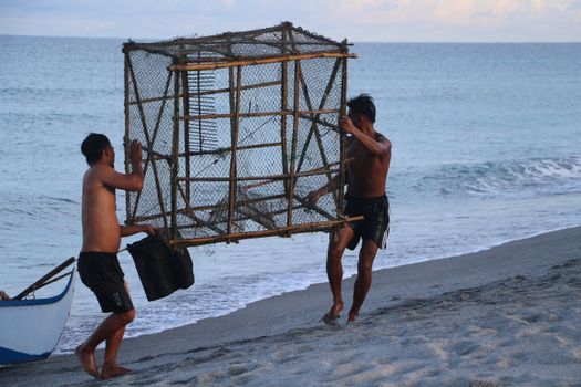 Two shirtless fishermen carrying a large fish trap on a sandy beach by the sea at sunset.