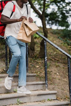 A courier carrying paper bags and wearing a red backpack walks down stairs outdoors.