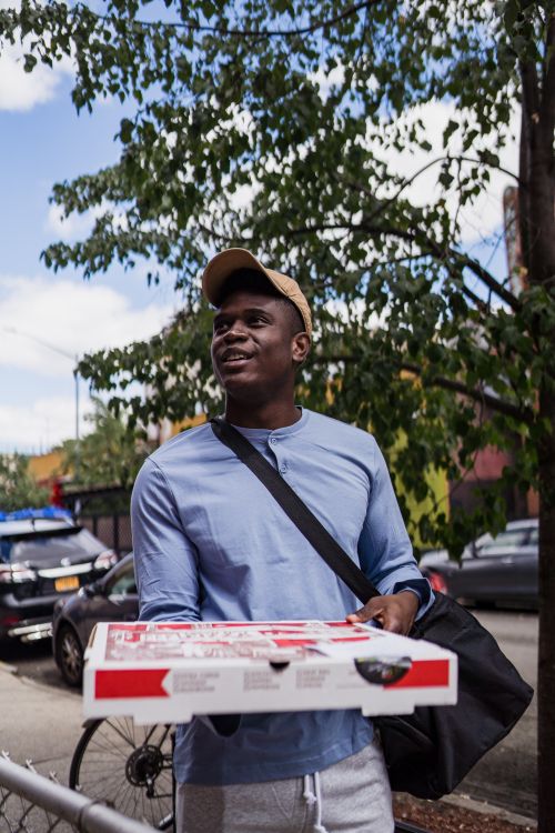 Free Portrait of deliveryman with pizza box walking on a city street, capturing the urban lifestyle. Stock Photo