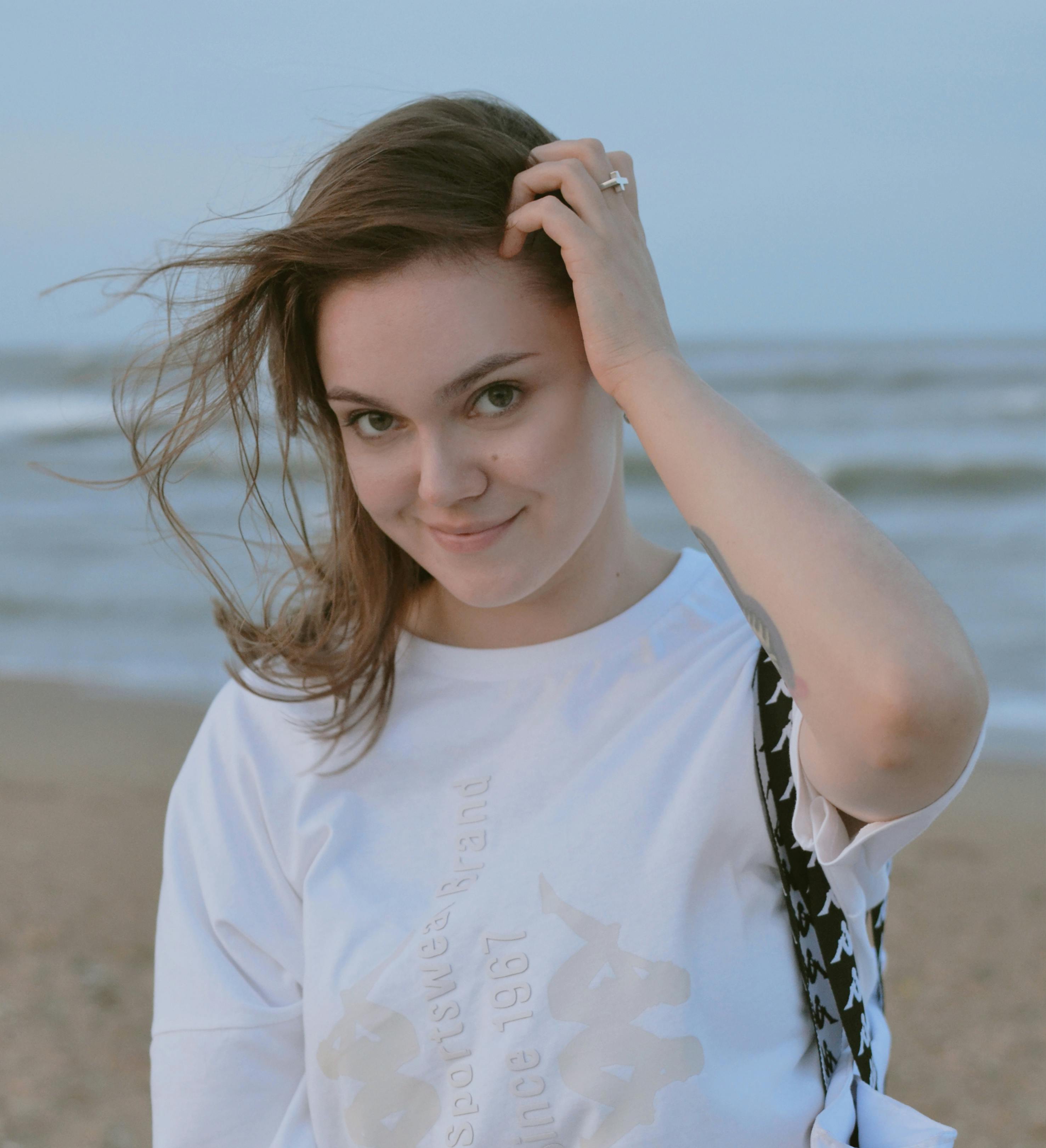 Portrait of a smiling young woman at the beach with wind-swept hair and casual attire.