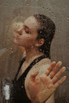 A woman with wet hair poses behind a glass pane covered in water droplets, creating a moody atmosphere.