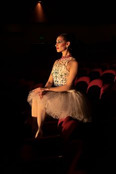 A poised ballerina in an elegant tutu sits gracefully in a dimly lit theater, exuding artistry and grace.