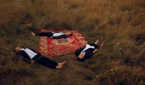 Free Three adults in suits lying on a rug in a meadow, photographed from above for a serene fashion concept. Stock Photo