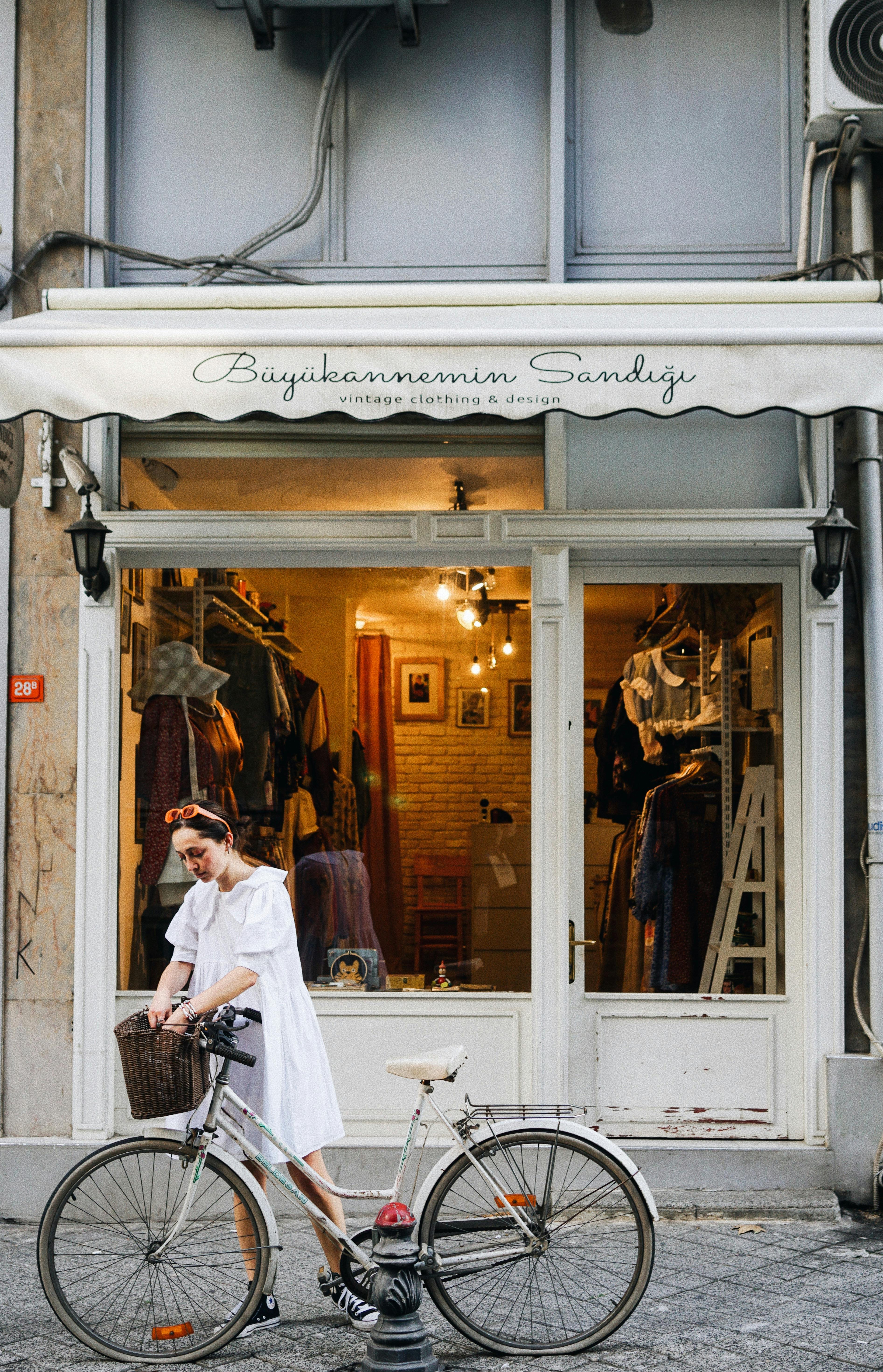 A woman in a white dress stands beside her bicycle in front of a vintage clothing shop.