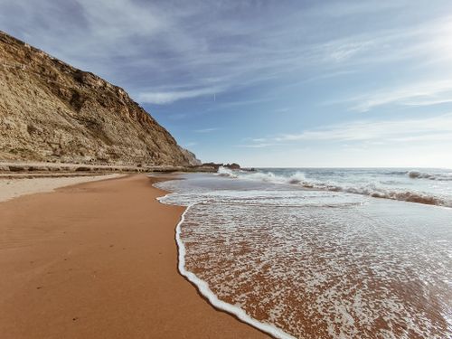 Free Scenic coastal view with waves, sand, and rocky cliff under a clear sky. Stock Photo