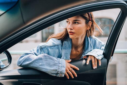 Stylish woman in denim jacket leaning on car door, exuding relaxed elegance.