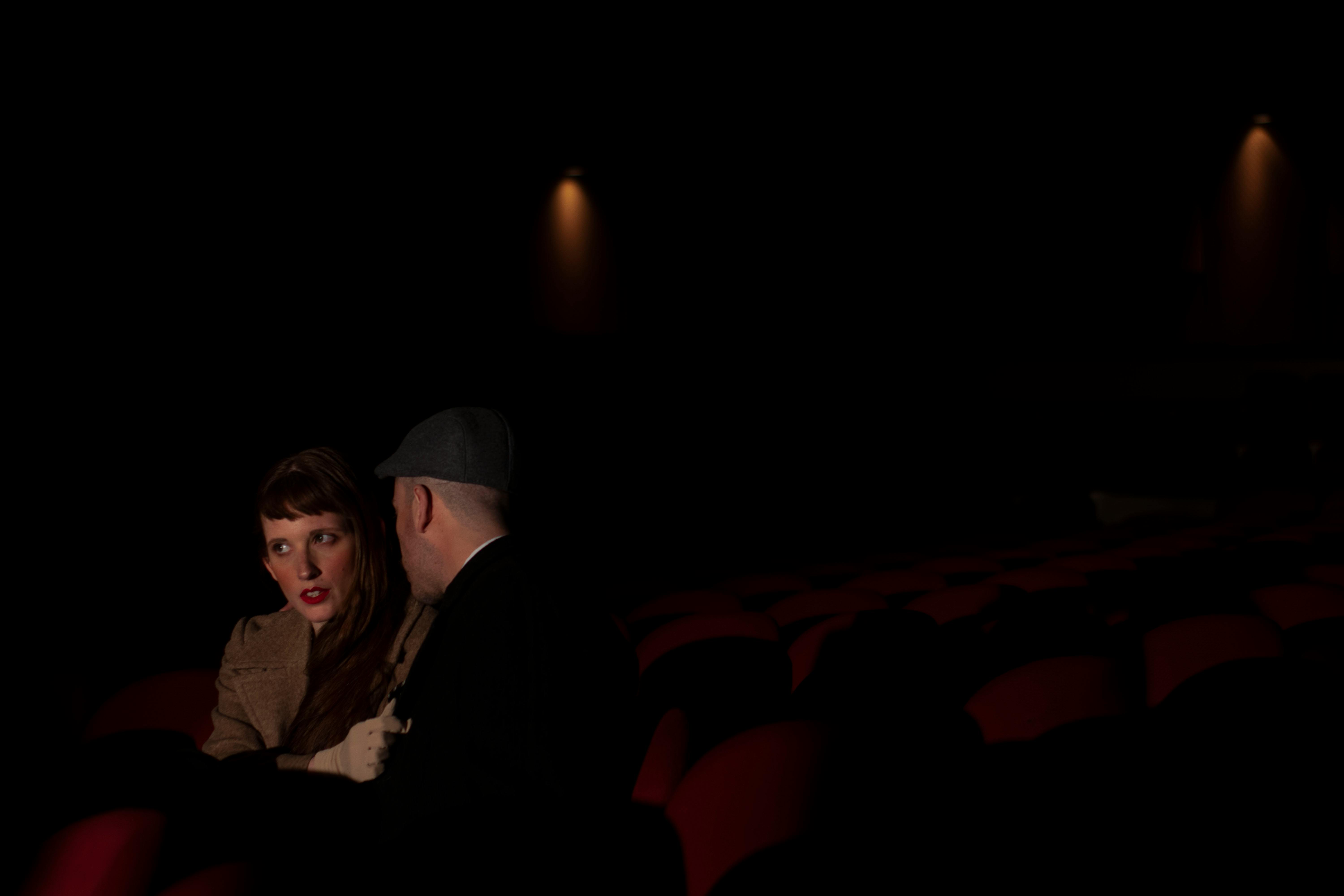Free A couple sitting closely in a dimly lit movie theater, whispering to each other. Stock Photo