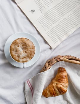 Cup of frothy coffee and freshly baked croissant on a bed with a magazine. Perfect morning breakfast scene.