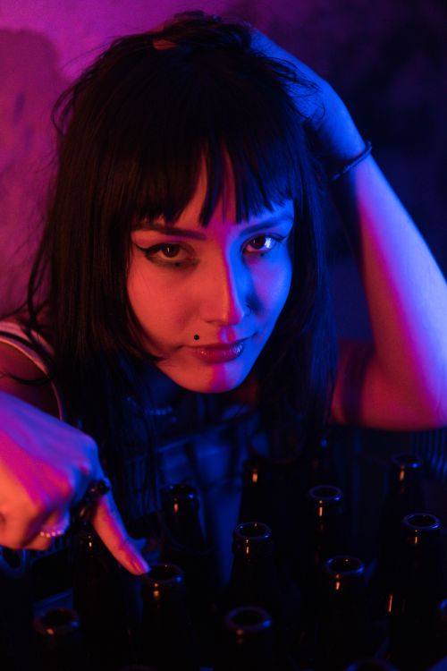 Free A woman with tattoos poses dramatically amid neon lights and glass bottles. Stock Photo