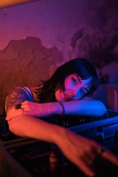A young woman with tattoos poses thoughtfully under cool and vibrant lighting in a nightclub.