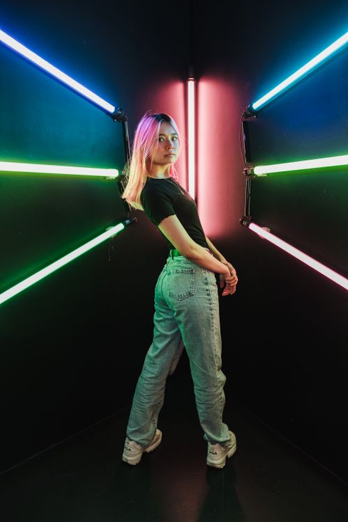 Free A young woman poses confidently in a neon-lit studio, featuring vibrant pink and green lights, showcasing modern style. Stock Photo