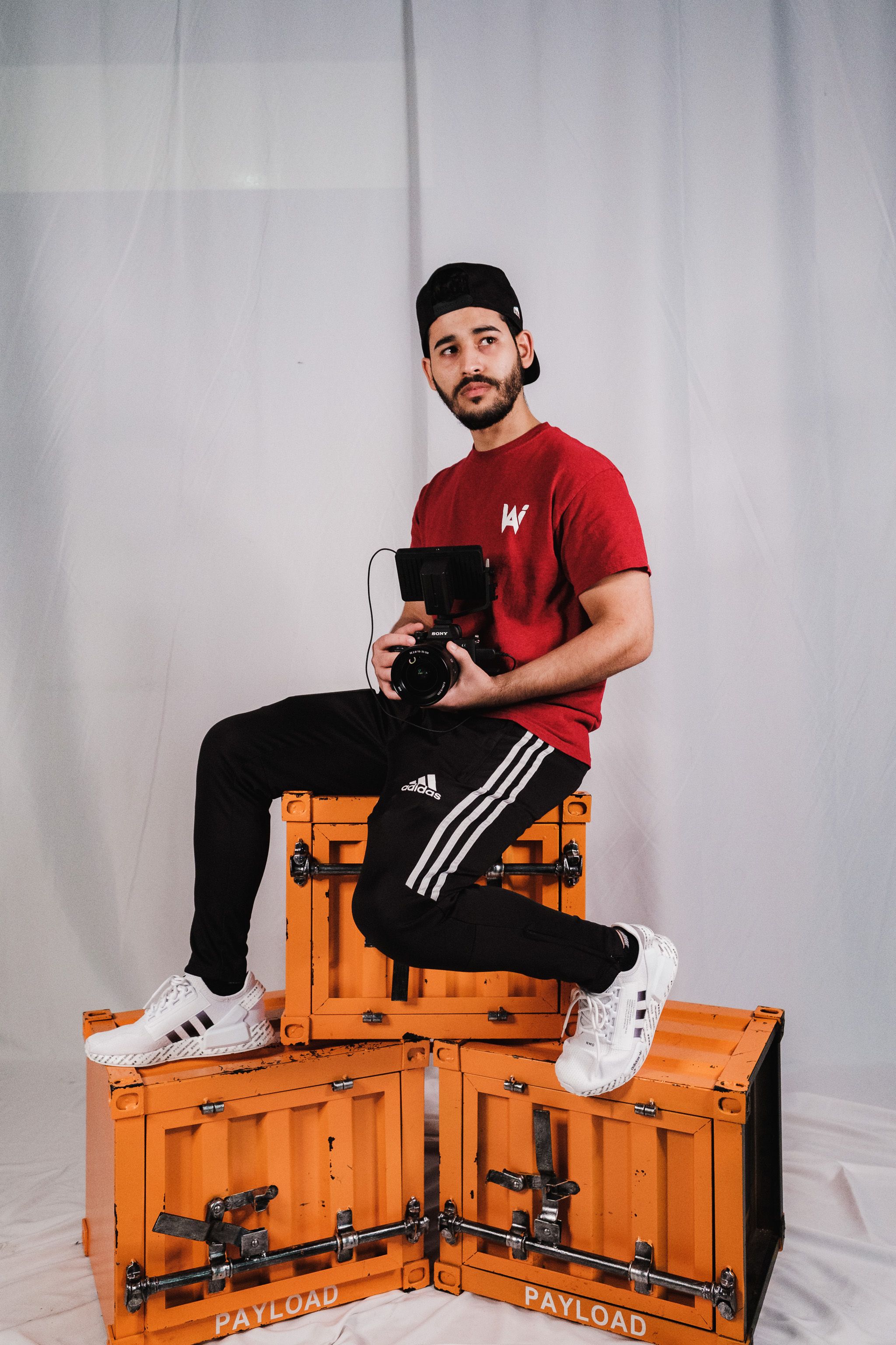 Man in red shirt holding camera sitting on orange boxes in studio.