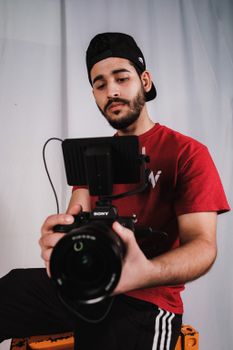 Young man with beard holding a professional camera, dressed in red indoors, looking focused.