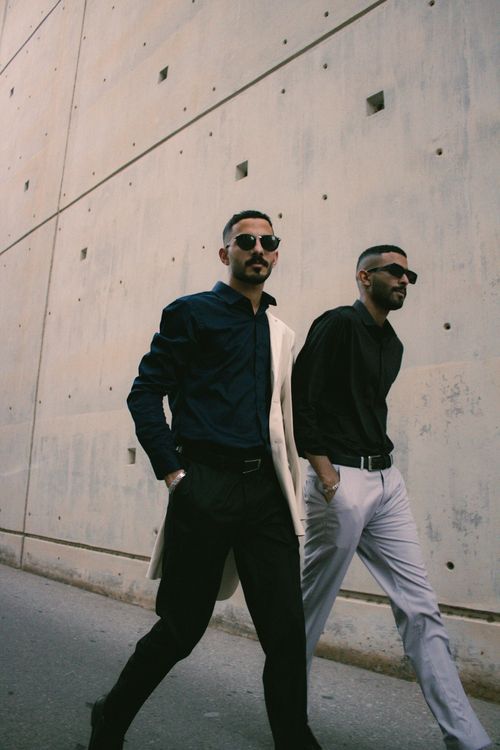 Free Two stylish men in black shirts walking past a modern wall in Muscat during the day. Stock Photo