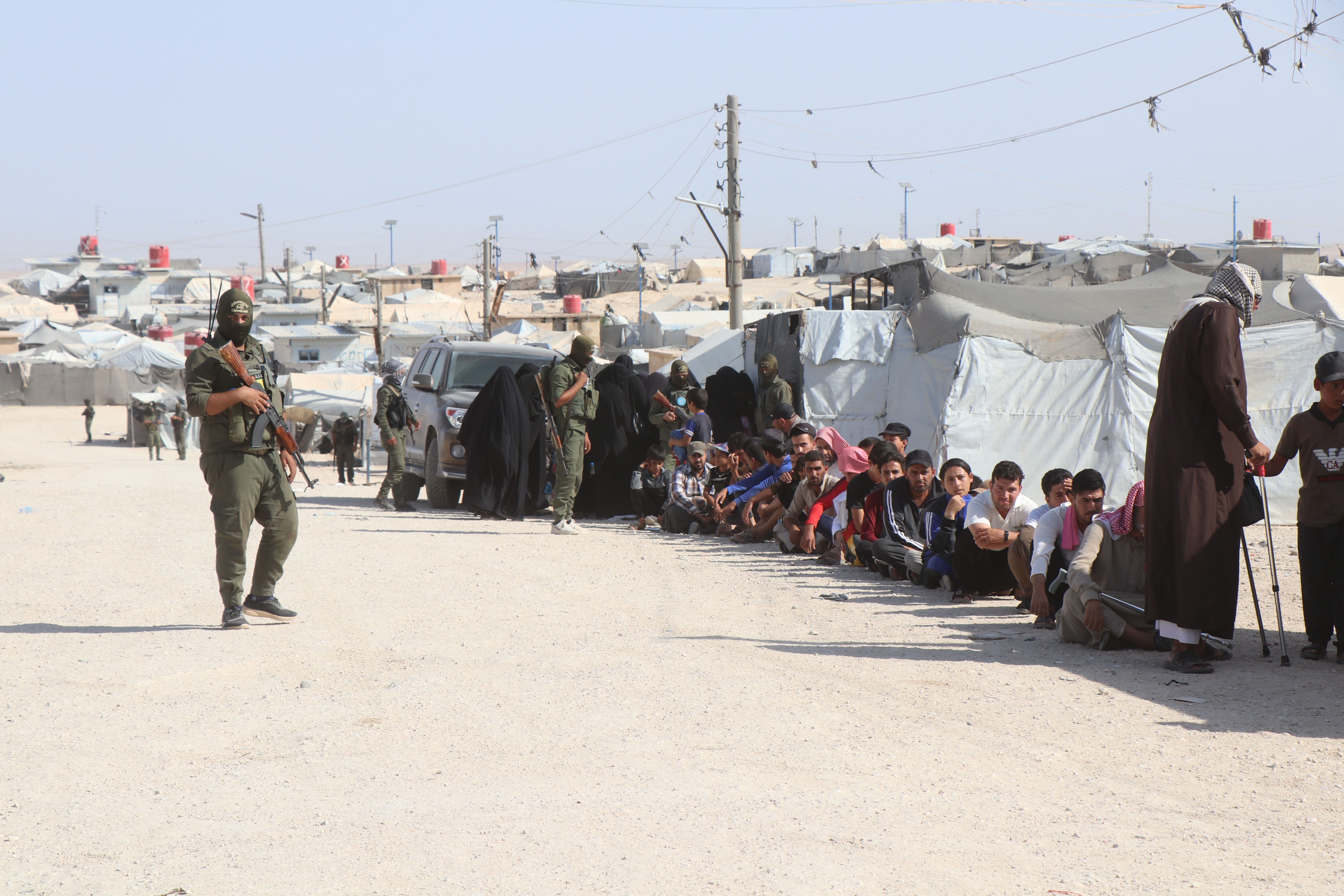 People queue beside tents in a refugee camp while armed guards patrol a dusty road